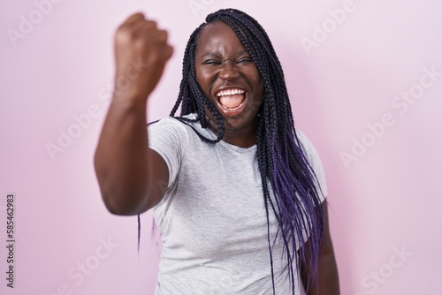Young african woman standing over pink background angry and mad raising fist frustrated and furious while shouting with anger. rage and aggressive concept.