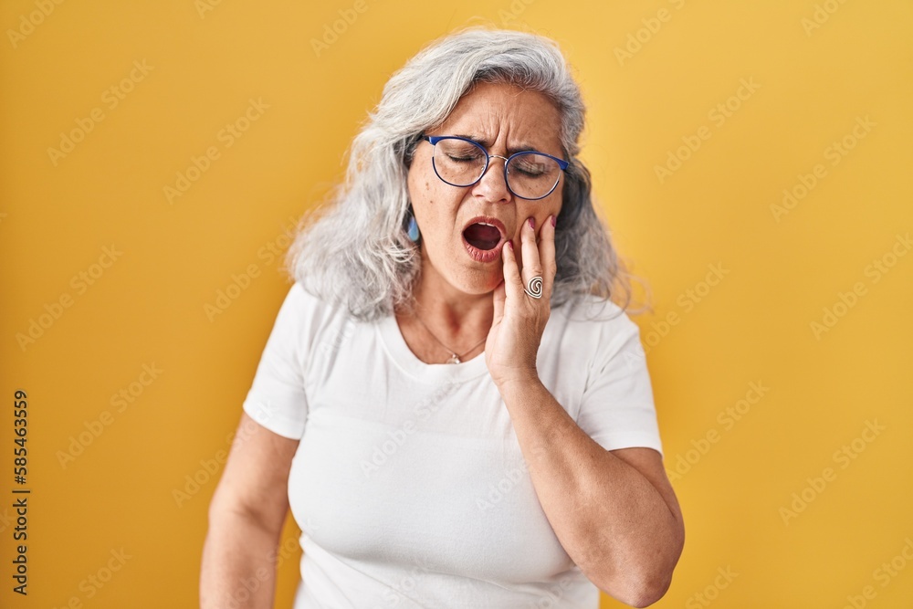 Middle age woman with grey hair standing over yellow background touching mouth with hand with painful expression because of toothache or dental illness on teeth. dentist