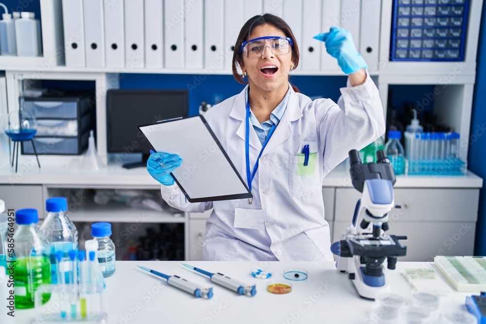 Hispanic young woman working at scientist laboratory angry and mad raising fist frustrated and furious while shouting with anger. rage and aggressive concept.