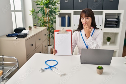 Young brunette doctor woman holding clipboard covering mouth with hand, shocked and afraid for mistake. surprised expression © Krakenimages.com
