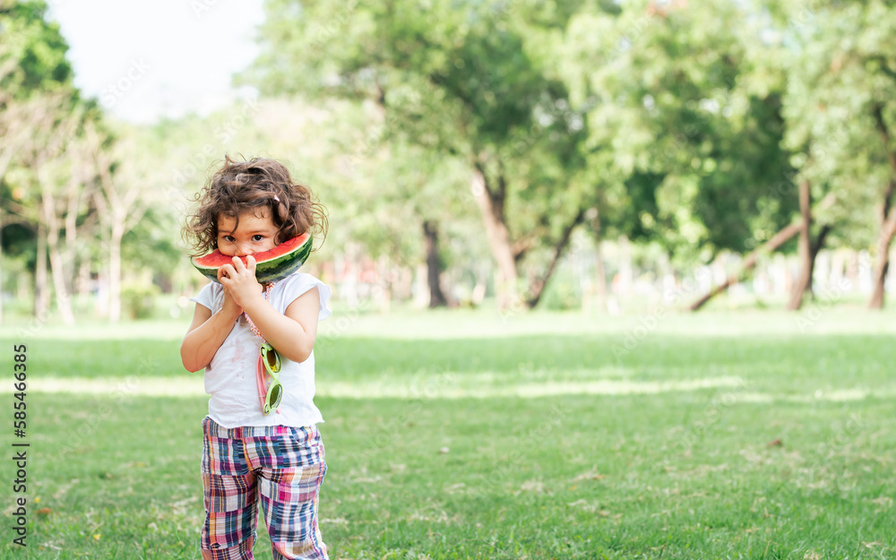 Caucasian little cute girl sitting, playing in outdoor green park for picnic, carrying, eating, biting fruit, watermelon with freshness. Education, Nature, Food Concept. Caucasian little cute girl sitting, playing in outdoor green park for picnic, carrying, eating, biting fruit, watermelon with freshness. Education, Nature, Food Concept.