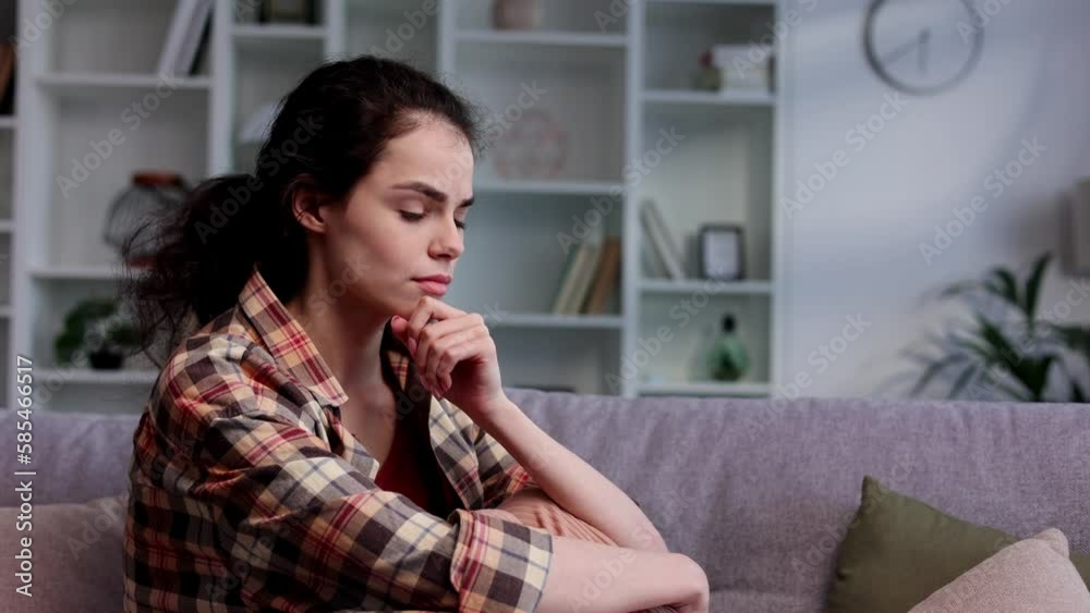 Young girl is deep in sad thoughts, thinks, looks away, feels tense and unhappy. Brunnete woman sitting on the couch have problen in personal life, feel depressed and broken.