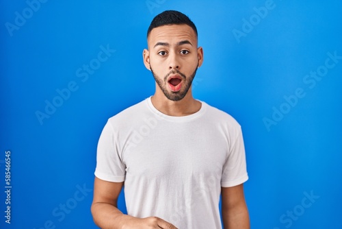 Young hispanic man standing over blue background afraid and shocked with surprise and amazed expression, fear and excited face.