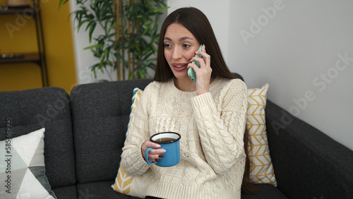 Young beautiful hispanic woman talking on smartphone drinking coffee at home