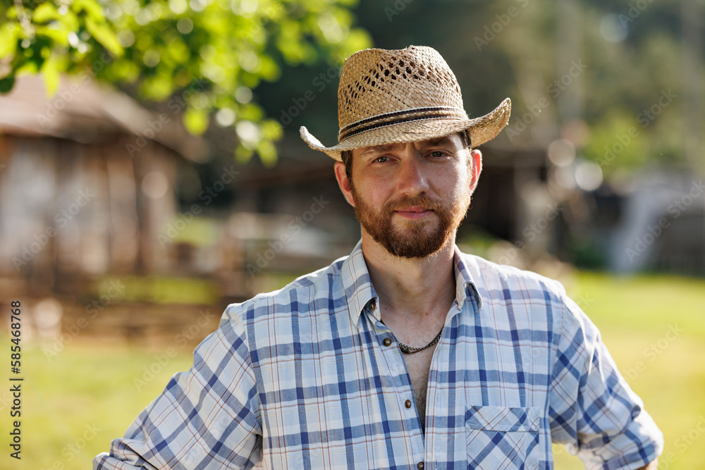Portrait of young Caucasian handsome bearded male farmer smiling to camera. Happy rancherman in straw hat at the eco farm. Industrial production of goat milk dairy products. Cattle farming