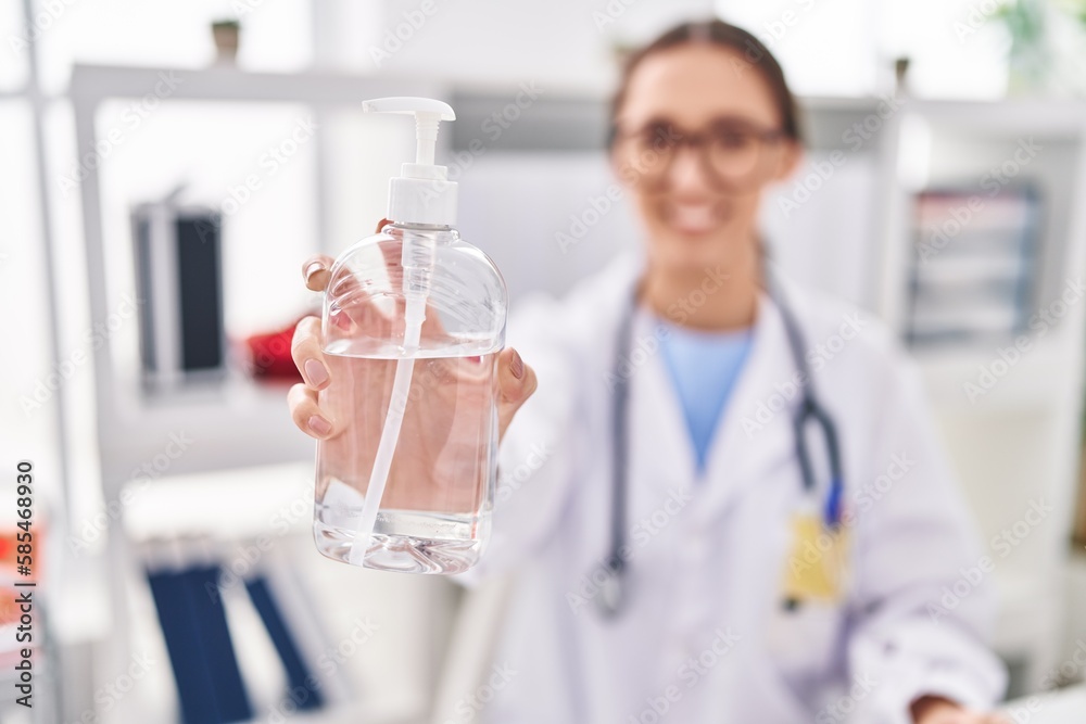 Young beautiful hispanic woman doctor holding sanitizer gel hands bottle at clinic Young beautiful hispanic woman doctor holding sanitizer gel hands bottle at clinic