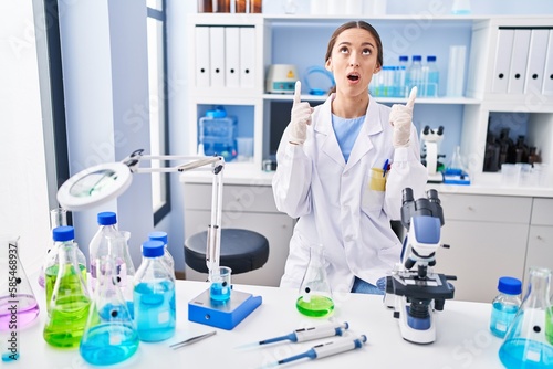 Young brunette woman working at scientist laboratory amazed and surprised looking up and pointing with fingers and raised arms.