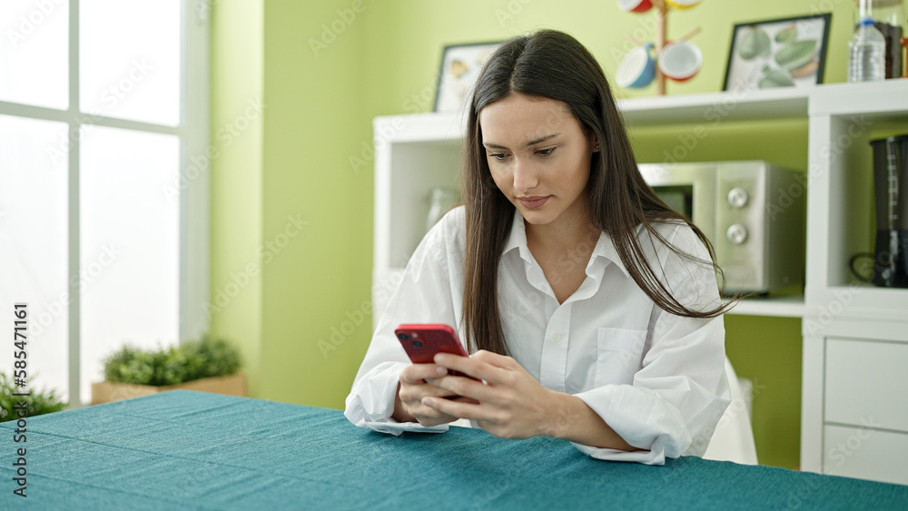 Young beautiful hispanic woman using smartphone sitting on table at dinning room Young beautiful hispanic woman using smartphone sitting on table at dinning room