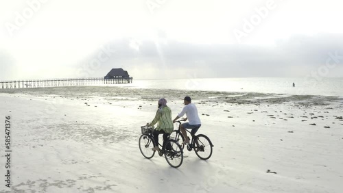 4K video couple of people on bicycles going along sandy beach at sunset with pier in background. Environment, healthy lifestyle, spending time outdoors concept. Zanzibar, Tanzania.