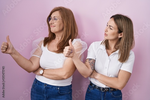 Hispanic mother and daughter wearing casual white t shirt over pink background looking proud, smiling doing thumbs up gesture to the side