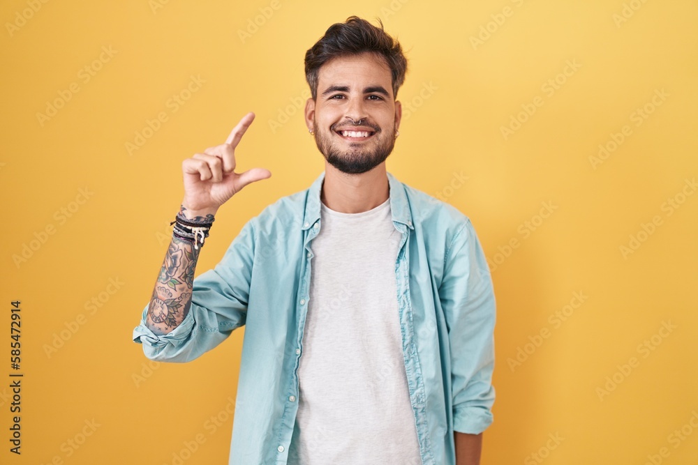 Young hispanic man with tattoos standing over yellow background smiling and confident gesturing with hand doing small size sign with fingers looking and the camera. measure concept. Young hispanic man with tattoos standing over yellow background smiling and confident gesturing with hand doing small size sign with fingers looking and the camera. measure concept.