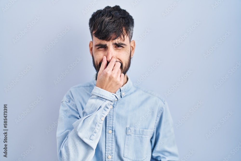 Young hispanic man with beard standing over blue background smelling something stinky and disgusting, intolerable smell, holding breath with fingers on nose. bad smell Young hispanic man with beard standing over blue background smelling something stinky and disgusting, intolerable smell, holding breath with fingers on nose. bad smell