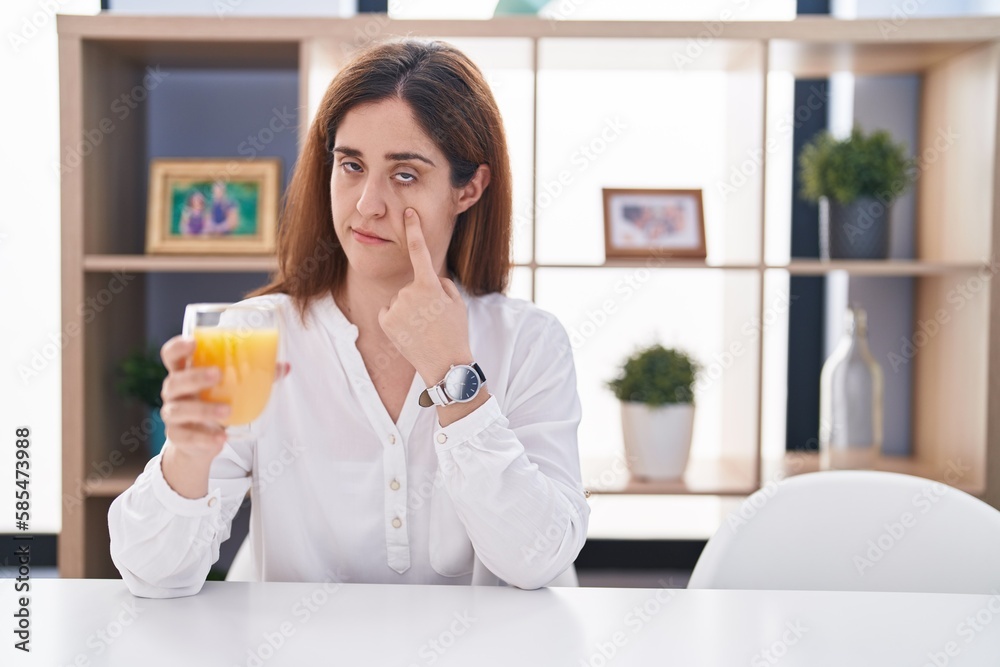 Brunette woman drinking glass of orange juice pointing to the eye watching you gesture, suspicious expression Brunette woman drinking glass of orange juice pointing to the eye watching you gesture, suspicious expression