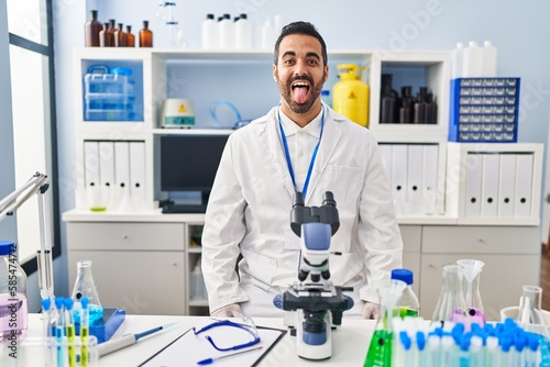 Young hispanic man with beard working at scientist laboratory sticking tongue out happy with funny expression. emotion concept.