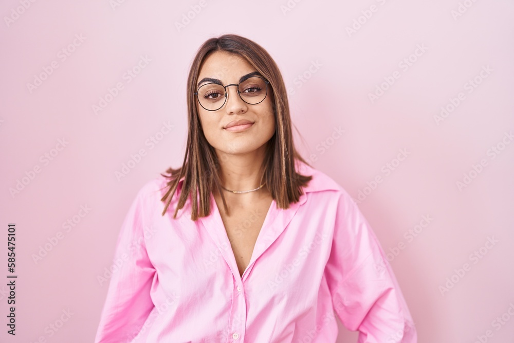 Young hispanic woman wearing glasses standing over pink background relaxed with serious expression on face. simple and natural looking at the camera. Young hispanic woman wearing glasses standing over pink background relaxed with serious expression on face. simple and natural looking at the camera.