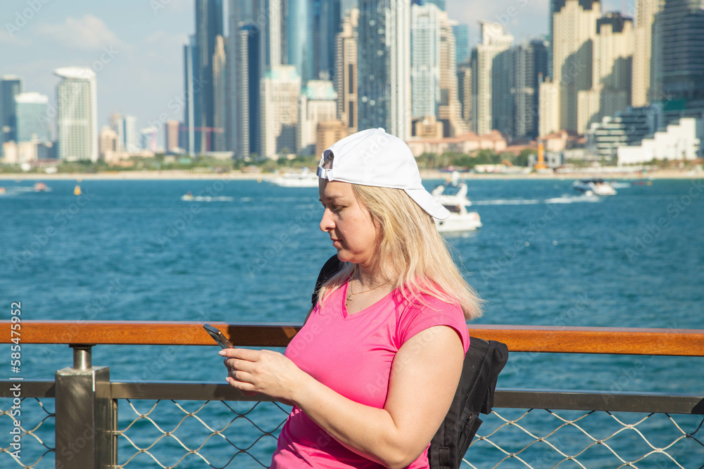 With a stunning perspective of the sea and modern buildings in Dubai, this portrait of a blonde tourist girl with a rucksack is taken while standing on the embankment.