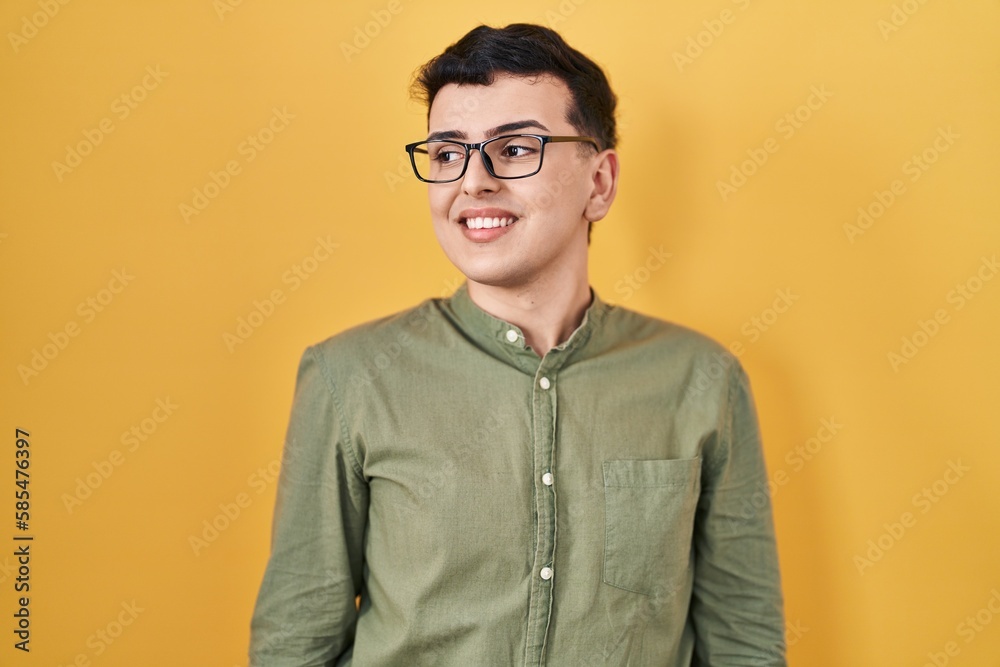 Fototapeta premium Non binary person standing over yellow background looking away to side with smile on face, natural expression. laughing confident.