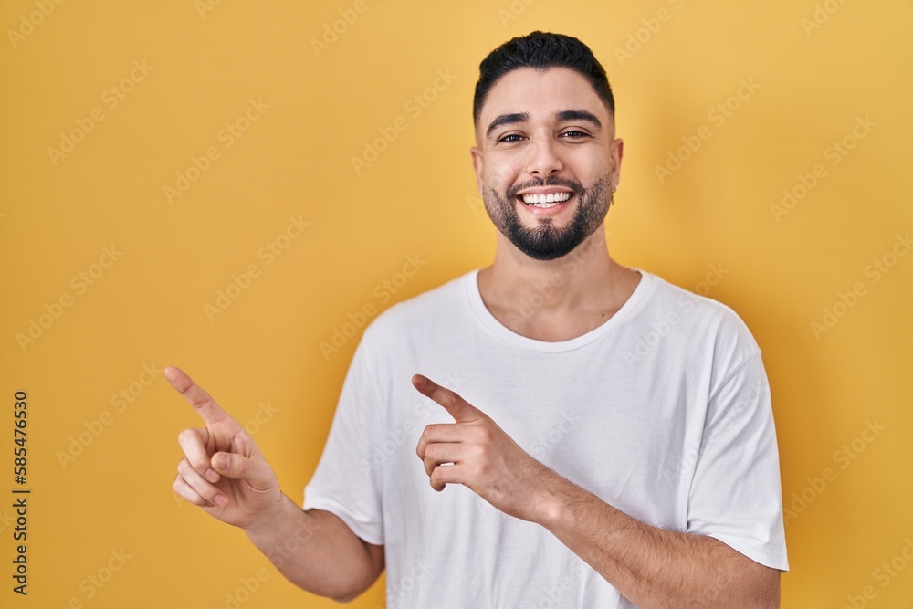Young handsome man wearing casual t shirt over yellow background smiling and looking at the camera pointing with two hands and fingers to the side. Young handsome man wearing casual t shirt over yellow background smiling and looking at the camera pointing with two hands and fingers to the side.