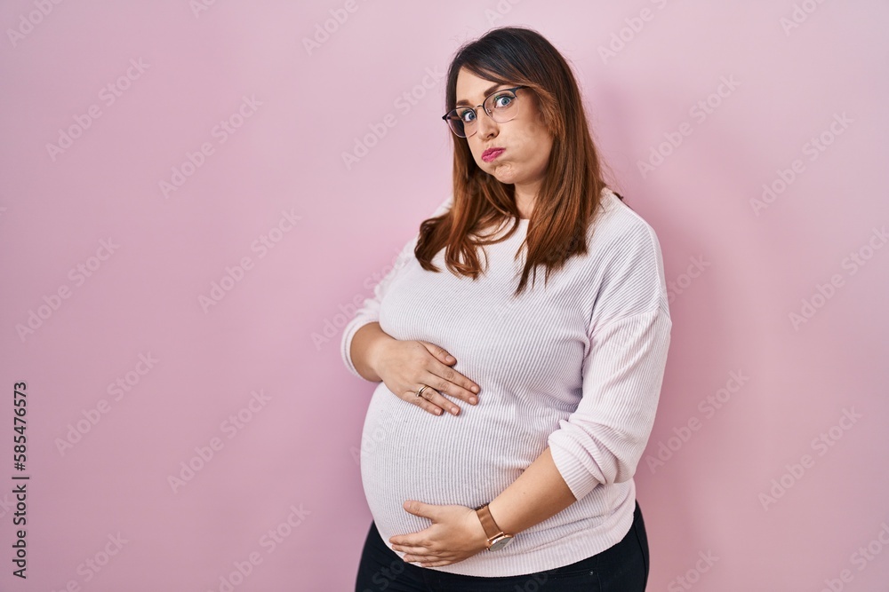 Pregnant woman standing over pink background puffing cheeks with funny face. mouth inflated with air, crazy expression. - obrazy, fototapety, plakaty 