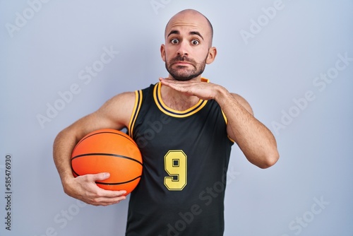 Young bald man with beard wearing basketball uniform holding ball cutting throat with hand as knife, threaten aggression with furious violence