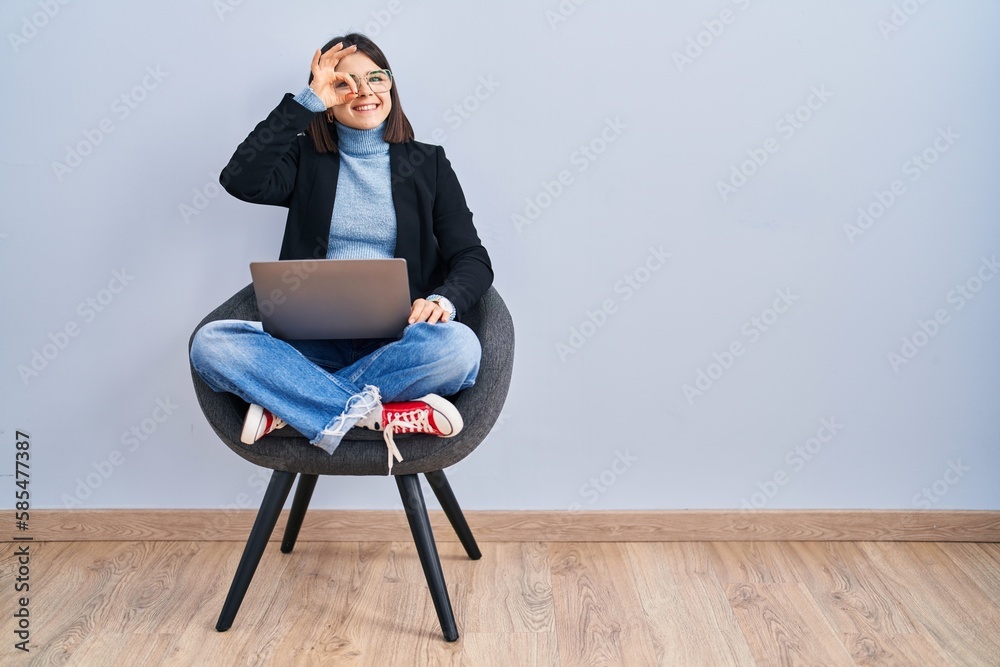 Young hispanic woman sitting on chair using computer laptop doing ok gesture with hand smiling, eye looking through fingers with happy face. Young hispanic woman sitting on chair using computer laptop doing ok gesture with hand smiling, eye looking through fingers with happy face.
