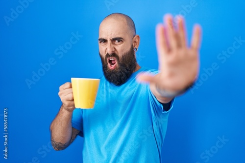 Young hispanic man with beard and tattoos drinking a cup of coffee doing stop gesture with hands palms, angry and frustration expression © Krakenimages.com