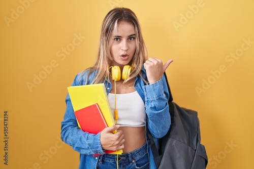 Young blonde woman wearing student backpack and holding books surprised pointing with hand finger to the side, open mouth amazed expression.