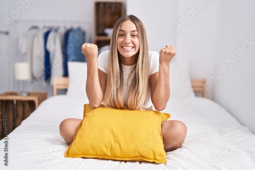 Young blonde woman sitting on the bed with pillow at home celebrating surprised and amazed for success with arms raised and open eyes. winner concept.