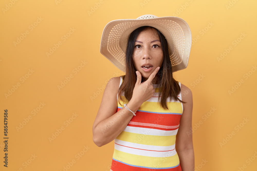 Middle age chinese woman wearing summer hat over yellow background looking fascinated with disbelief, surprise and amazed expression with hands on chin Middle age chinese woman wearing summer hat over yellow background looking fascinated with disbelief, surprise and amazed expression with hands on chin