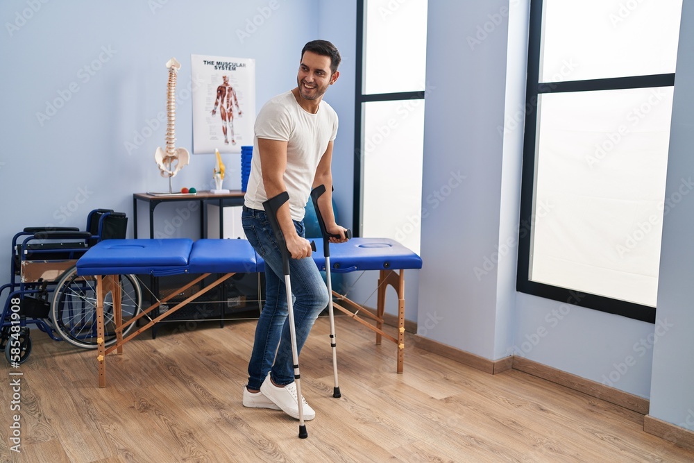 Fototapeta premium Young hispanic man injured physiotherapy patient using crutches walking at rehab clinic