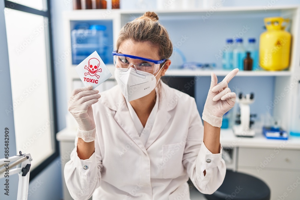Young woman working at scientist laboratory holding toxic banner smiling happy pointing with hand and finger to the side Young woman working at scientist laboratory holding toxic banner smiling happy pointing with hand and finger to the side