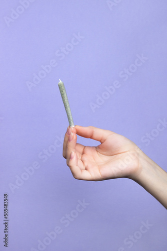 A woman's hand with natural nails holds a joint with medical marijuana on a light purple background.  copy space
