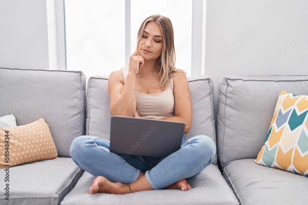 Blonde caucasian woman using laptop at home sitting on the sofa serious face thinking about question with hand on chin, thoughtful about confusing idea Blonde caucasian woman using laptop at home sitting on the sofa serious face thinking about question with hand on chin, thoughtful about confusing idea