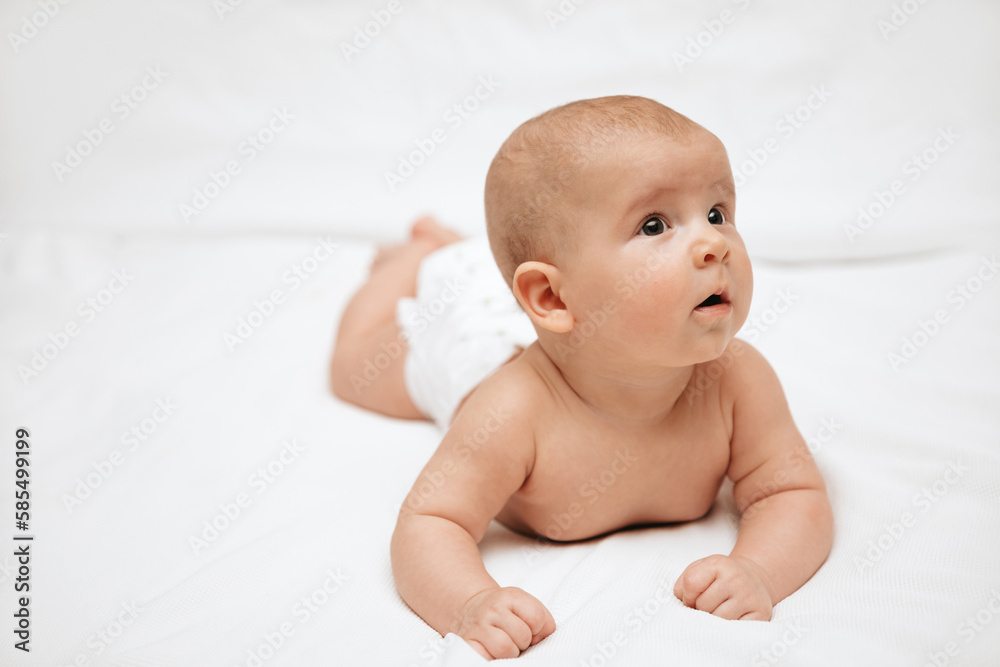 A newborn baby in a diaper is lying on stomach on a white background and looking to the side. A newborn baby in a diaper is lying on stomach on a white background and looking to the side.