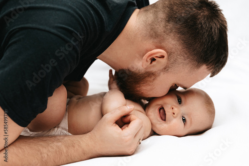 Dad is kissing newborn daughter on a white blanket. The baby is lying with daddy on the bed and laughing.