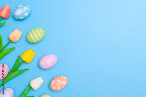 Happy Easter holiday greeting card design concept. Colorful Easter Eggs and spring flowers on blue background. Flat lay, top view, copy space.