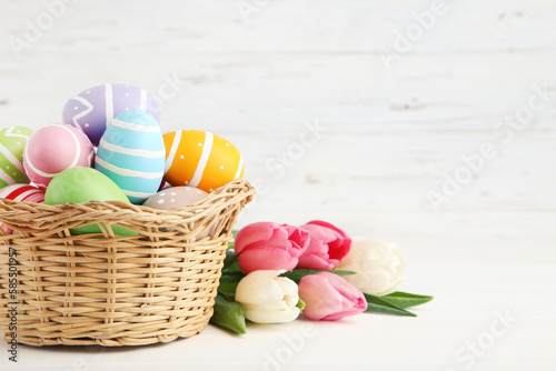 Colorful easter eggs in basket and bouquet of tulips on white wooden background