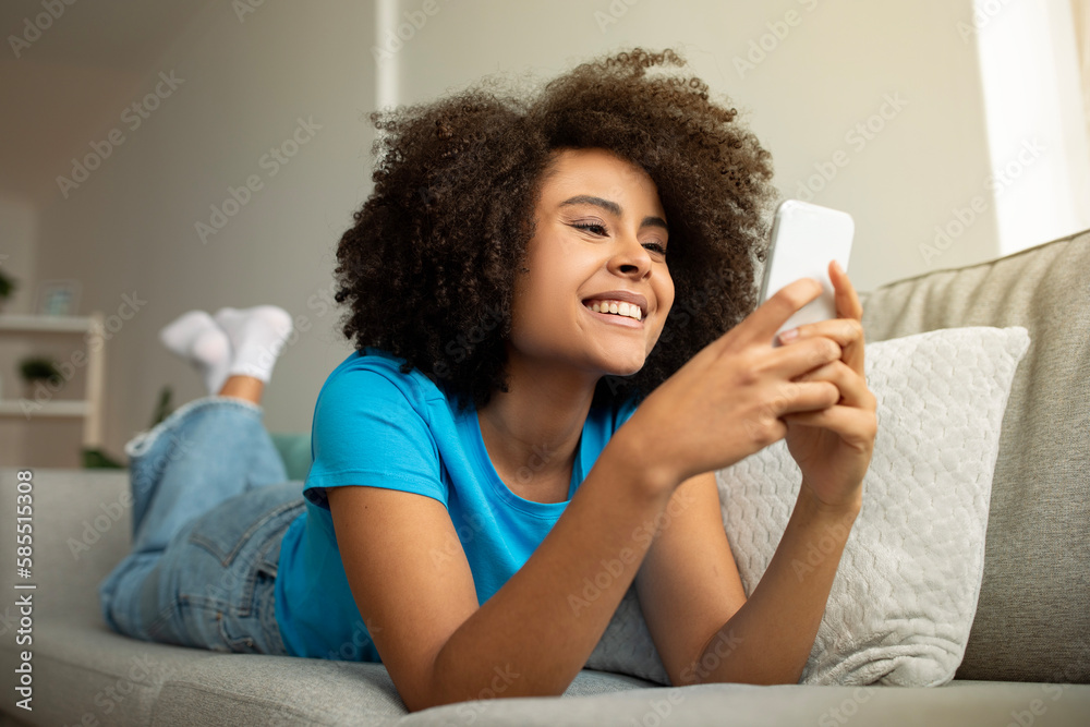 Cheerful young curly african american lady typing on smartphone, enjoy social networks, chat, lies on sofa Cheerful young curly african american lady typing on smartphone, enjoy social networks, chat, lies on sofa
