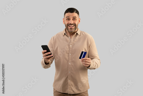 Excited middle aged man shopping online with mobile phone and credit card, looking at camera with excitement