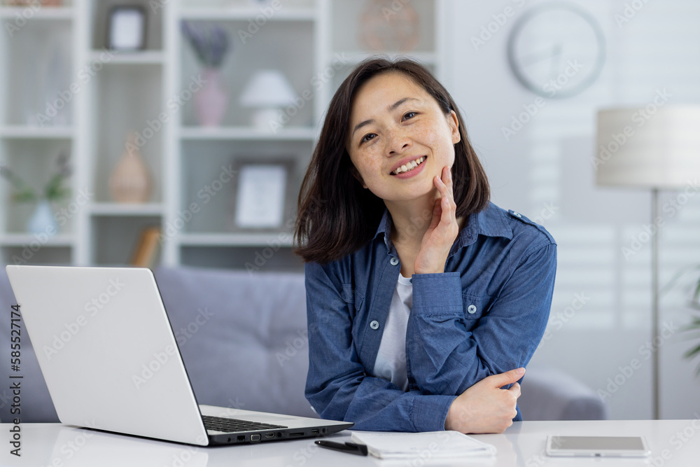 Portrait of young beautiful asian woman at home, woman smiling and looking at camera, working remotely in home office using laptop while sitting at table in living room. Portrait of young beautiful asian woman at home, woman smiling and looking at camera, working remotely in home office using laptop while sitting at table in living room.