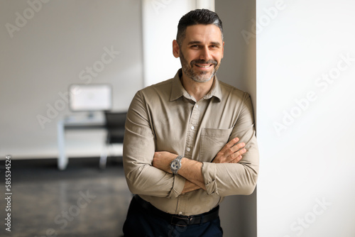 Portrait of confident middle aged businessman standing in office hall, posing with crossed hands and leaning on wall