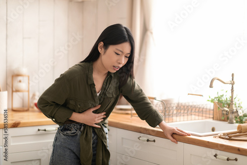 Young asian woman feeling sick in kitchen, touching her belly photo