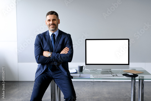 Confident middle aged businessman leaning on table, posing with folded arms near computer with blank screen, mockup