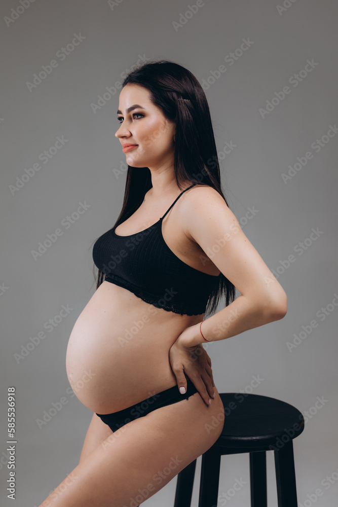 Fototapeta premium Portrait Of Happy Pregnant Woman In Lingerie With Smile Face Made Of Cream On Her Belly Posing Over Beige Background, Beautiful Young Expectant Female In Underwear Smiling At Camera.