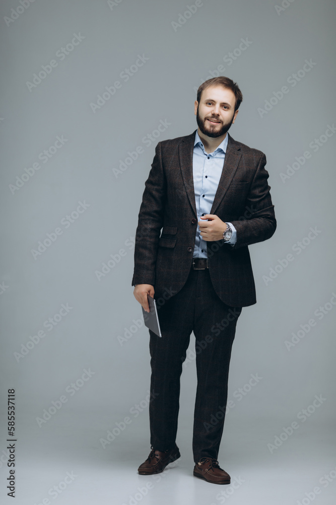 Attractive smiling white collar worker standing on grey background, looking at camera and holding tablet. Morning routine before work concept