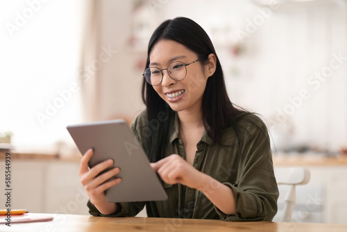 Positive young asian woman using digital tablet at kitchen