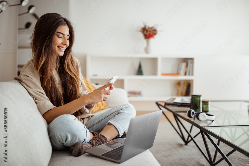Smiling millennial mixed race woman looks at phone, reads message, has meeting, work with laptop Smiling millennial mixed race woman looks at phone, reads message, has meeting, work with laptop