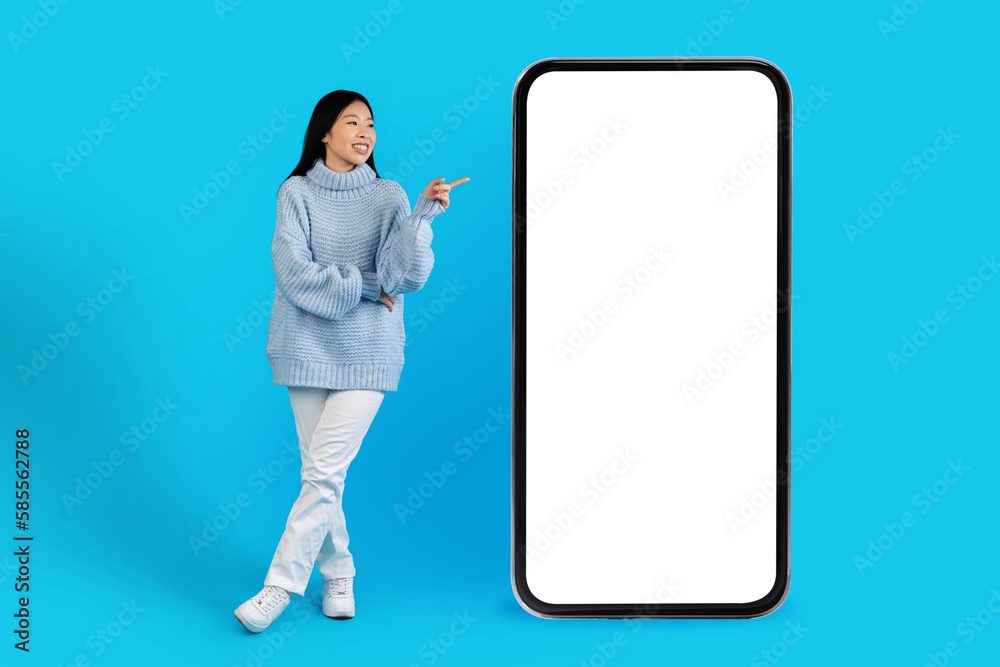 Cheerful asian woman in warm sweater pointing at smartphone, mockup Cheerful asian woman in warm sweater pointing at smartphone, mockup