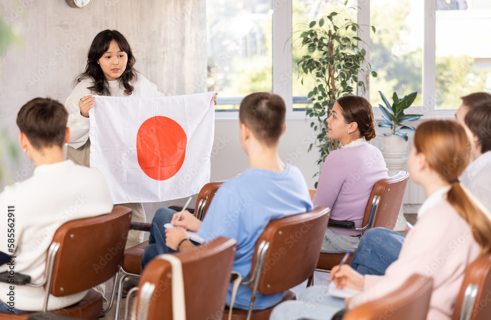 Young teacher showing big flag of Japan to group of grown-up student in light classroom
