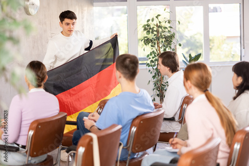 Young man tells his classmates the history of the state of Germany, holding the national flag of Germany in his hands